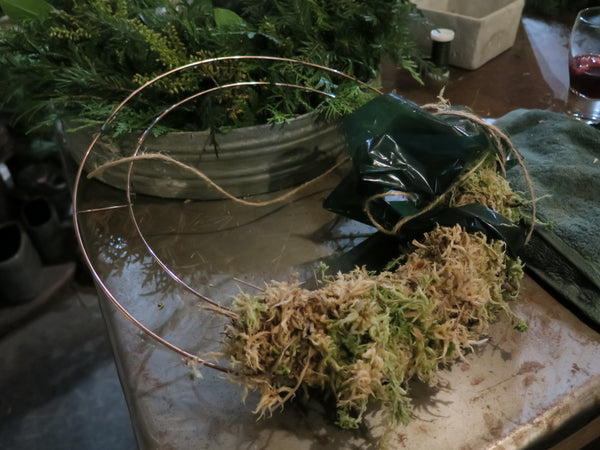 A wreath in progress lies on a table, crafted from a wire frame and partially adorned with moss. In the background, there's a container brimming with assorted greenery. A small glass filled with red liquid is visible on the table next to a green cloth, creating the perfect scene for an Event Wreath Workshop.