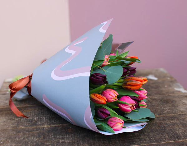 The Armful of Tulip Bouquet by Wix, a romantic gift, features vibrant pink, orange, and purple tulips wrapped with decorative paper and a brown ribbon. It rests elegantly on a wooden surface set against a light pink and beige background.