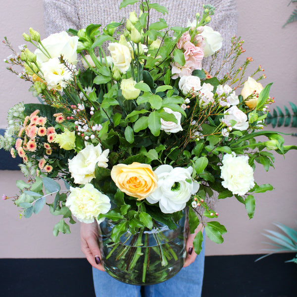 A person in a beige sweater and blue jeans holds the 'Jar of Joy' Vase Arrangement by Wild About Flowers, featuring pastel roses and green foliage, creating a luxurious floral gift against a light-colored wall.