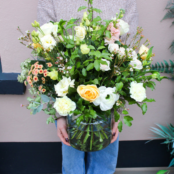 A person in a beige sweater and blue jeans holds the 'Jar of Joy' Vase Arrangement by Wild About Flowers, a luxury glass vase filled with assorted fresh blooms, standing before a pink wall.