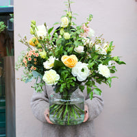 A person in a gray sweater holds Wild About Flowers' 'Jar of Joy' vase arrangement—a luxury mix of yellow, white, and pastel roses with greenery—making an elegant Valentine’s Day flower gift against a pale background.