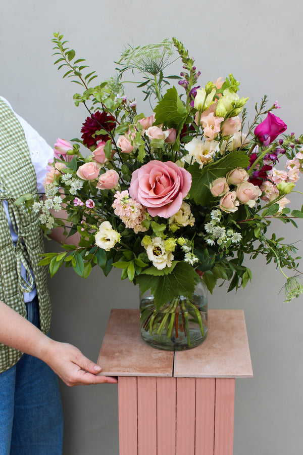 An individual showcases the "Juicy bunch" from Wild About Flowers, a wooden stand cradling a glass vase brimming with a seasonal bouquet in vibrant pink roses, alongside white and purple blooms with lush greenery, all set against a light gray backdrop. The person is dressed in a green checkered vest and blue jeans.