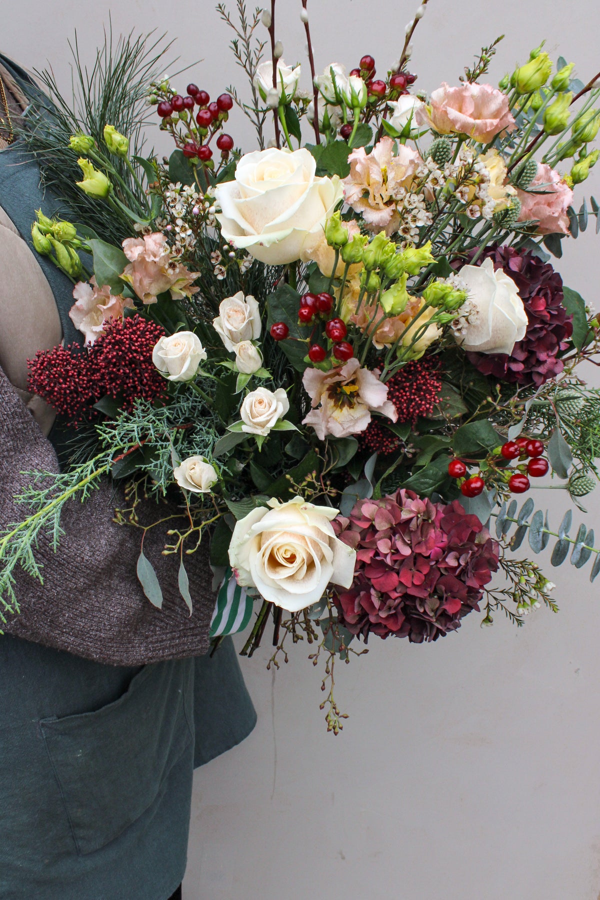 A person holds the Love Actually Hand Tied by Wild About Flowers—a festive mix of white roses, red hydrangeas, green foliage, and berries—against a plain background. Ideal for flower delivery in Keynsham.
