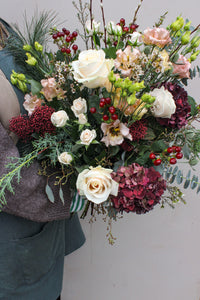 A person holds the Love Actually Hand Tied by Wild About Flowers—a festive mix of white roses, red hydrangeas, green foliage, and berries—against a plain background. Ideal for flower delivery in Keynsham.