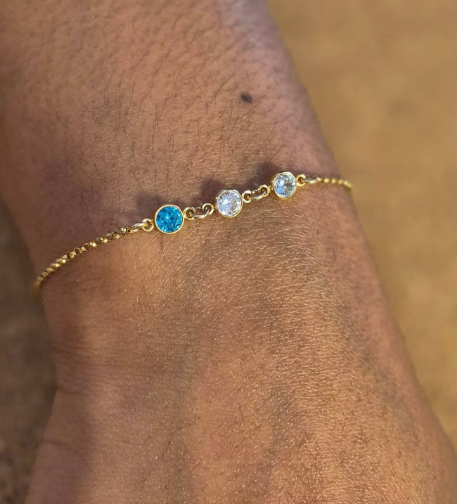 A close-up of Event's Summer Sessions gold bracelet, featuring one blue and two clear round gems, worn on a wrist as chic permanent jewellery against a blurred brown background.