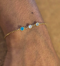 A close-up of Event's Summer Sessions gold bracelet, featuring one blue and two clear round gems, worn on a wrist as chic permanent jewellery against a blurred brown background.