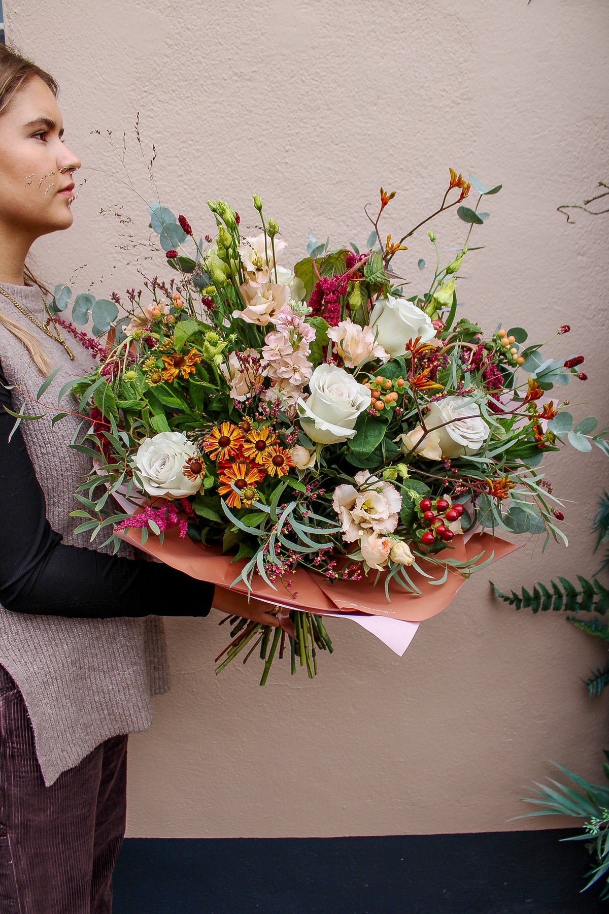 A person holds the "Maple Latte" bouquet by Wild About Flowers—an autumn-winter arrangement with white roses, orange and pink blooms, greenery, and berries—against a light-colored wall.