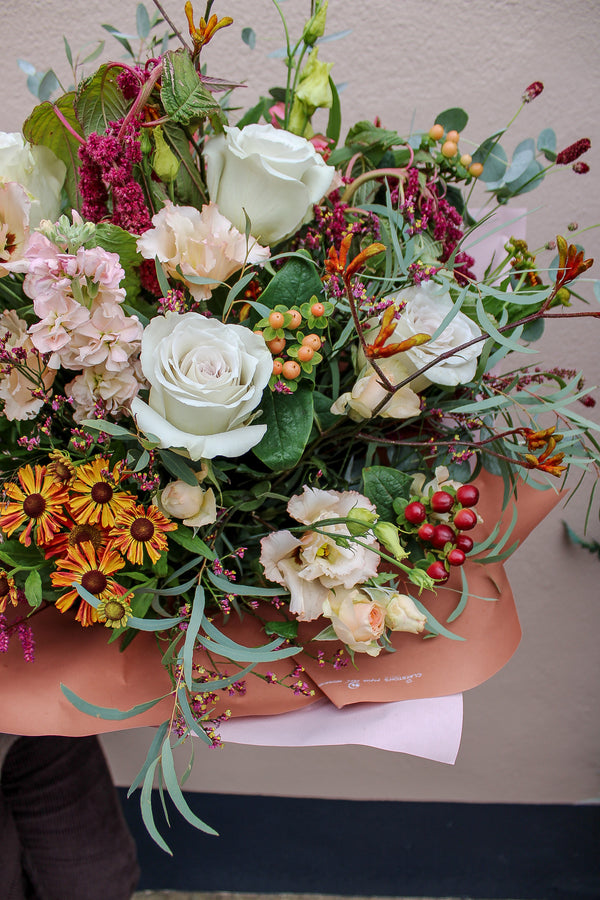 Maple Latte by Wild About Flowers is a vibrant hand-tied bouquet with white roses, yellow daisies, pink lisianthus, orange and red berries, and green foliage, wrapped in brown and pink paper—ideal for autumn-winter flower lovers.