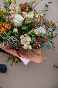 The Maple Latte by Wild About Flowers is a hand-tied bouquet with white roses, petite pink and orange autumn-winter blooms, red berries, and green foliage, all wrapped in peach and pink paper against a light beige background.