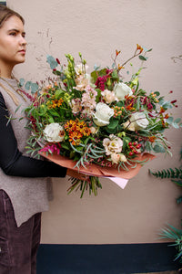 A person holds Wild About Flowers’ Maple Latte bouquet, a vibrant hand-tied mix of white roses, red berries, autumnal yellow and orange blooms, and green foliage against a neutral wall.