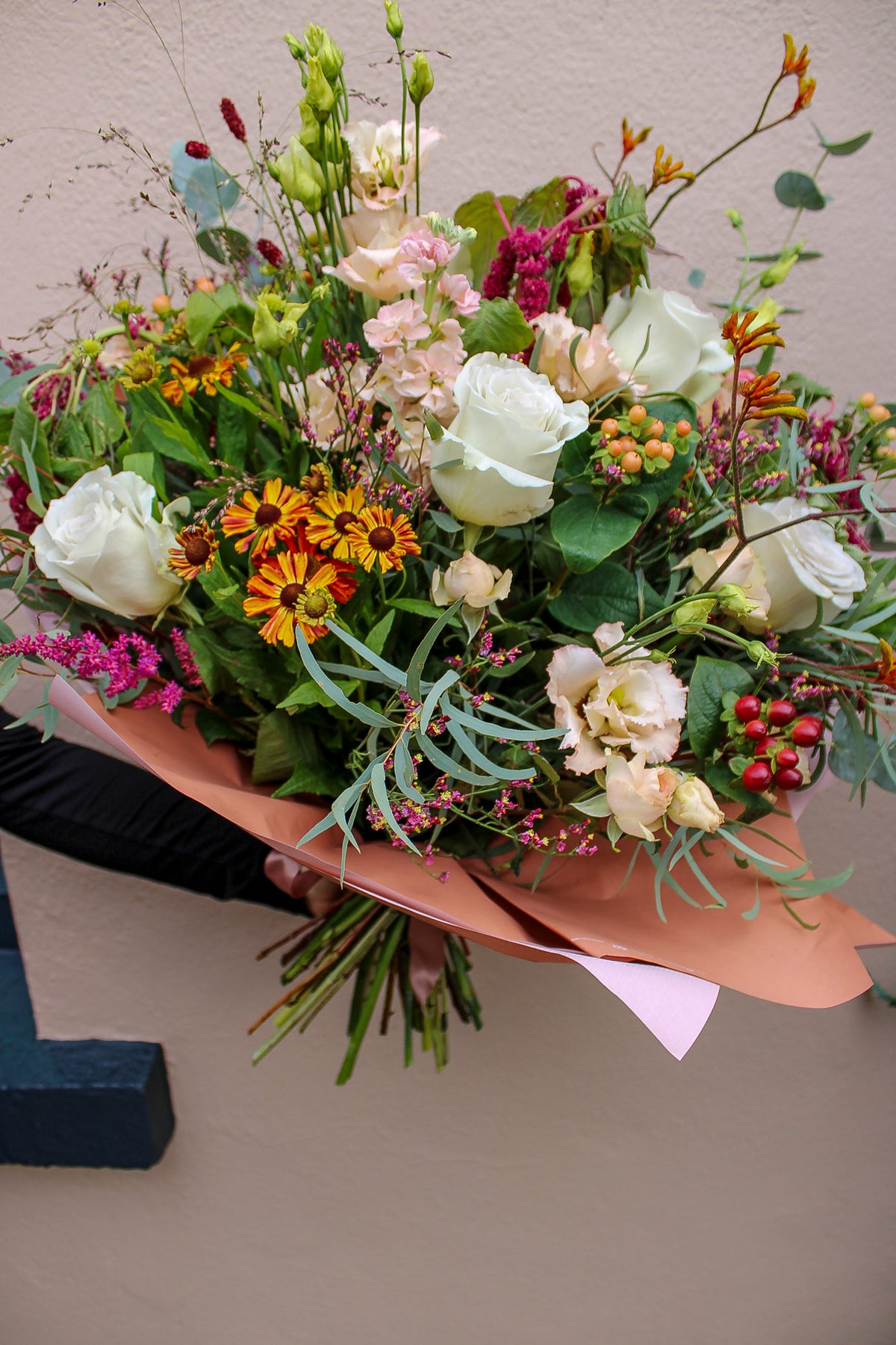 A hand holds the Maple Latte bouquet by Wild About Flowers—white roses, yellow daisies, pink blooms, greenery, and berries—hand-tied in eco paper against a beige wall, capturing the warmth of autumn-winter flowers.