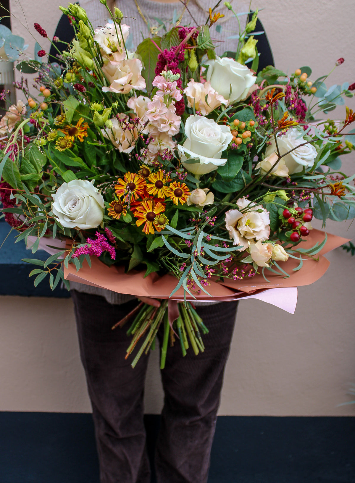 A person holds the Wild About Flowers "Maple Latte" bouquet, featuring hand-tied white roses, yellow, orange, and pink blooms with mixed greenery in brown paper. The cozy autumn-winter arrangement creates a seasonal vibe; their face isn’t visible.