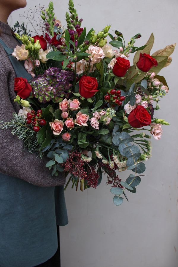 A person in a blue shirt and gray sweater holds the Wild About Flowers Mulled Wine Hand Tied, featuring red roses, pink seasonal blooms, berries, eucalyptus, and mixed greenery against a plain light background.