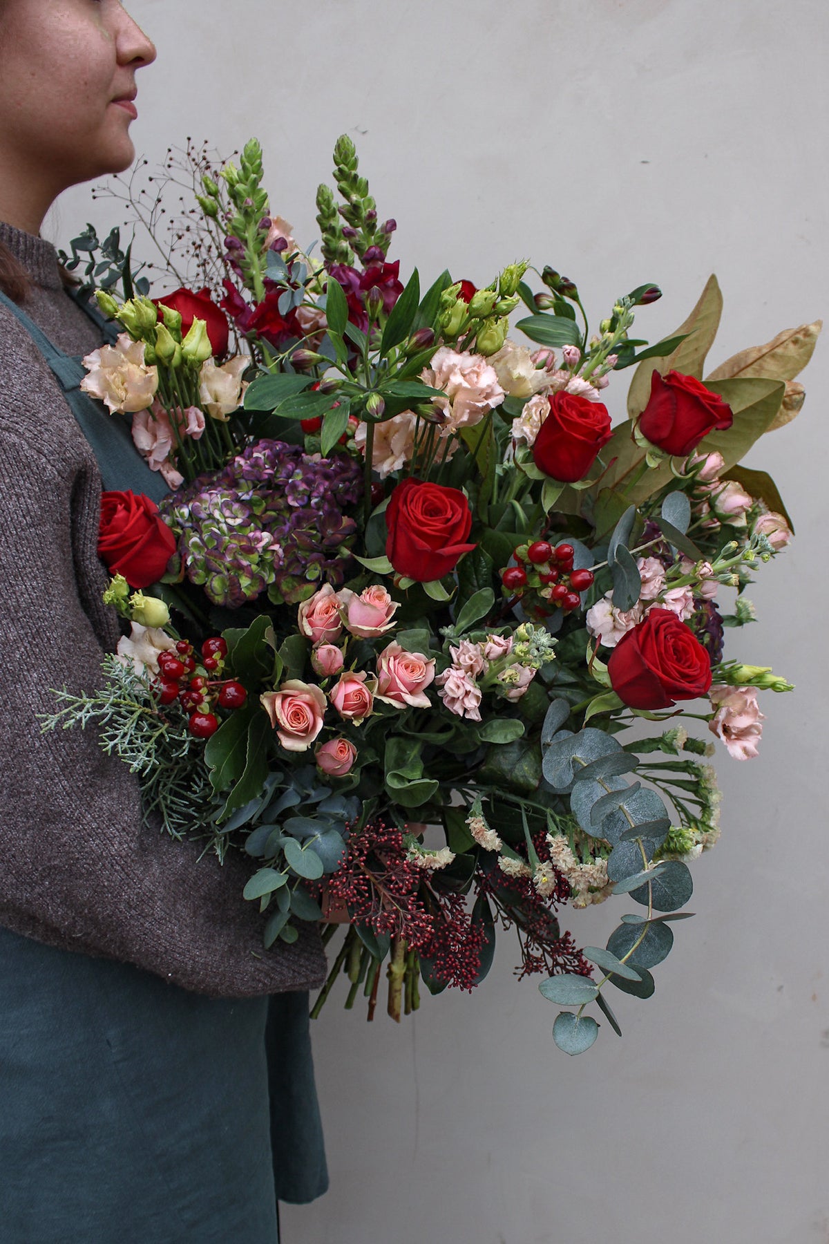 A person in a knit sweater and apron holds the Wild About Flowers Mulled Wine Hand Tied bouquet, featuring vibrant red roses, pink and purple winter blooms, berries, and greenery against a plain light background.