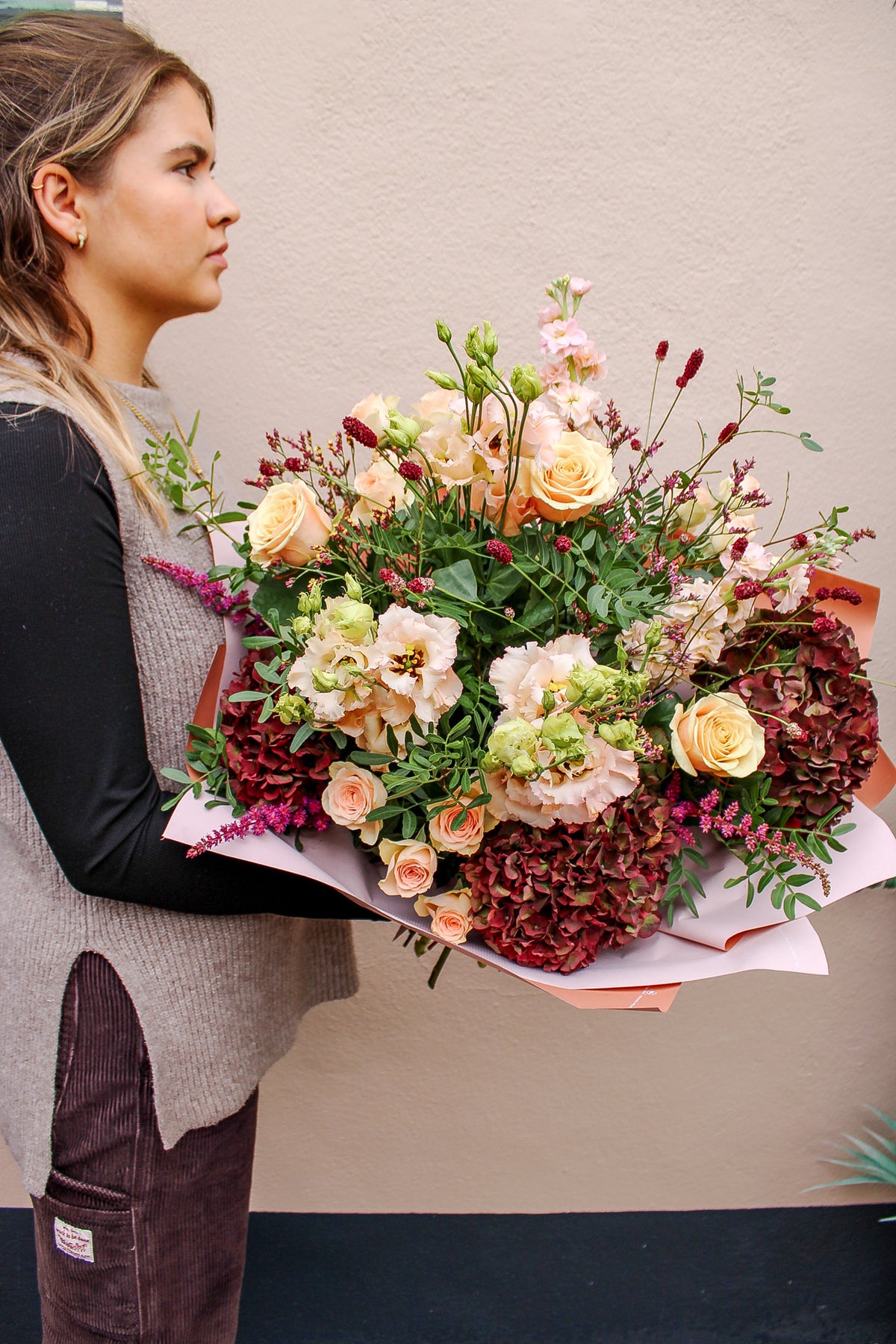 A woman with light brown hair stands in profile holding the Wild About Flowers Pecan Pie bouquet—featuring roses and hydrangeas in pink paper—against a beige wall, perfect for an autumn flower delivery.