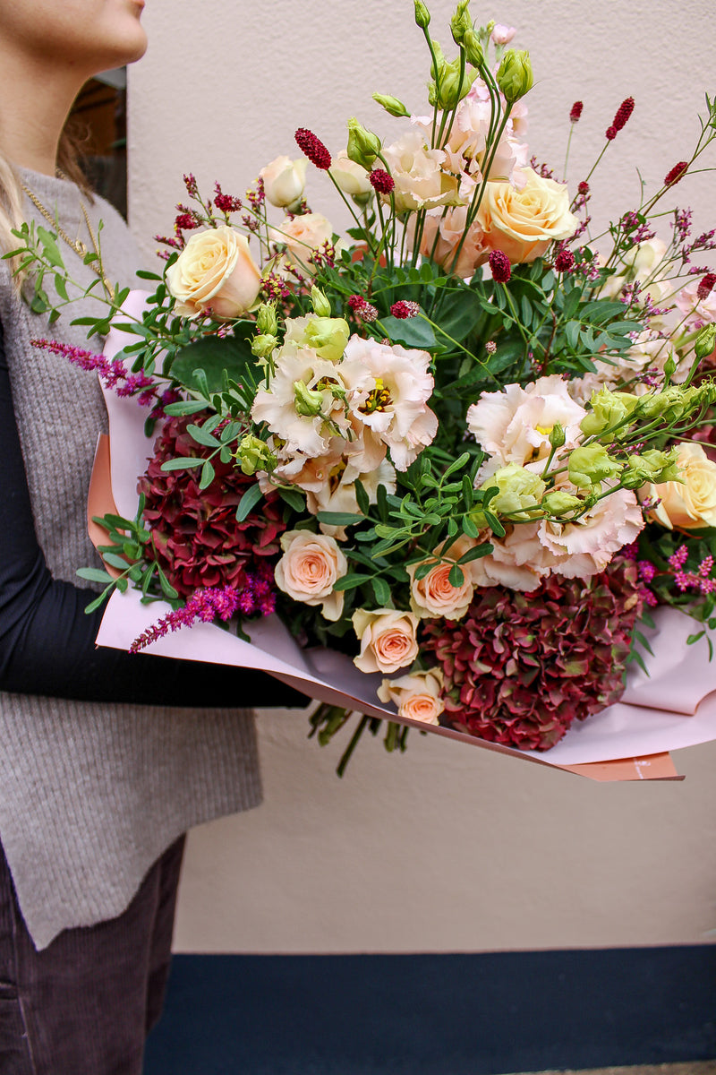A person holds the Wild About Flowers "Pecan Pie" bouquet, a large hand-tied arrangement with pink and cream roses, pale lisianthus, burgundy hydrangeas, and greenery, wrapped in pink paper against a plain light background.