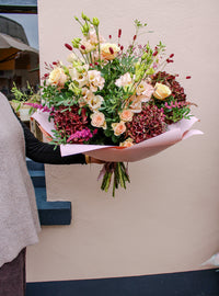 A person holds the Wild About Flowers "Pecan Pie" bouquet—pale peach roses, burgundy and pink blooms with green foliage—hand-tied and wrapped in light pink paper, set against a light-colored wall.