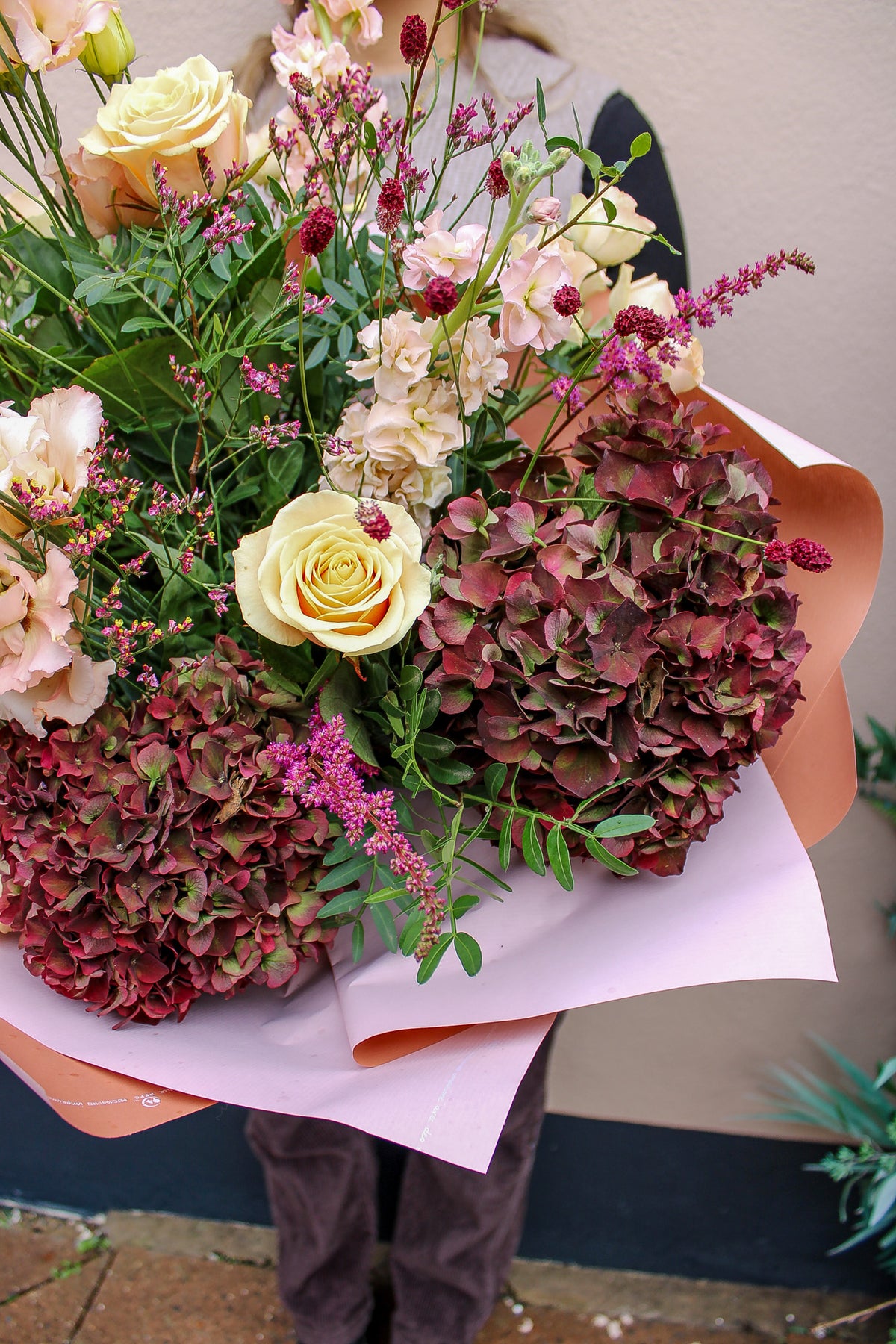 A person holds the Pecan Pie bouquet by Wild About Flowers, wrapped in pink paper with yellow roses, hydrangeas, greenery, and magenta blooms—perfect for autumn delivery from your local Keynsham shop. Their face is not visible.