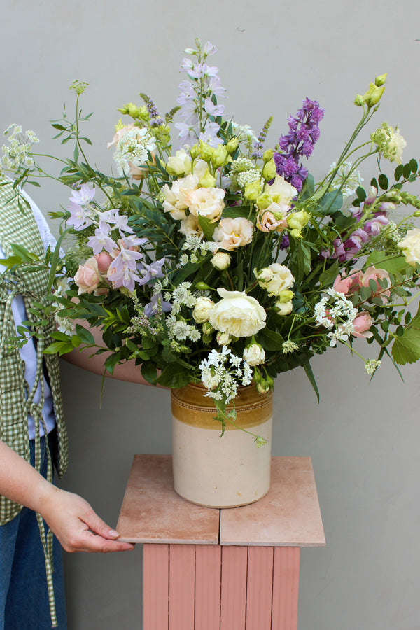 A person skillfully assembles a large floral bouquet in the Pretty Pot by Wild About Flowers, setting it on a pink pedestal. This breathtaking arrangement showcases roses, lisianthus, and greenery in shades of pink, white, and purple. A plain light gray wall serves as the backdrop, accentuating the elegance of this sustainable decor.