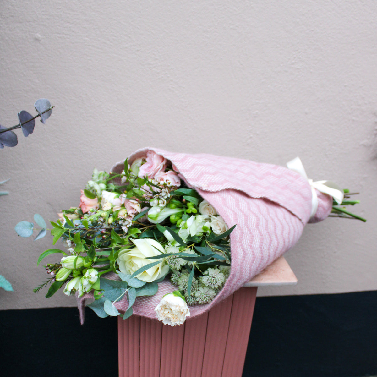 The ‘Strawberry Kisses’ Valentine’s Flower Wrap by Wild About Flowers features assorted blooms in a pink patterned wrap, displayed on a ribbed pink pedestal against a light beige wall—perfect for a romantic gift.