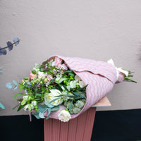 The ‘Strawberry Kisses’ Valentine’s Flower Wrap by Wild About Flowers features assorted blooms in a pink patterned wrap, displayed on a ribbed pink pedestal against a light beige wall—perfect for a romantic gift.
