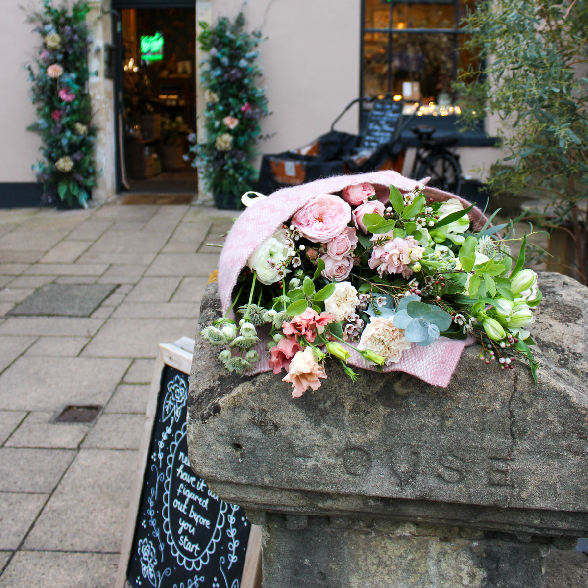 Wild About Flowers’ ‘Strawberry Kisses’ Valentine’s Flower Wrap of pink and white blooms sits on a stone post by a shop with a floral arch, chalkboard sign, and bicycle—hinting at local flower delivery for the perfect romantic gift.