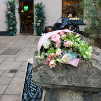 Wild About Flowers’ ‘Strawberry Kisses’ Valentine’s Flower Wrap of pink and white blooms sits on a stone post by a shop with a floral arch, chalkboard sign, and bicycle—hinting at local flower delivery for the perfect romantic gift.