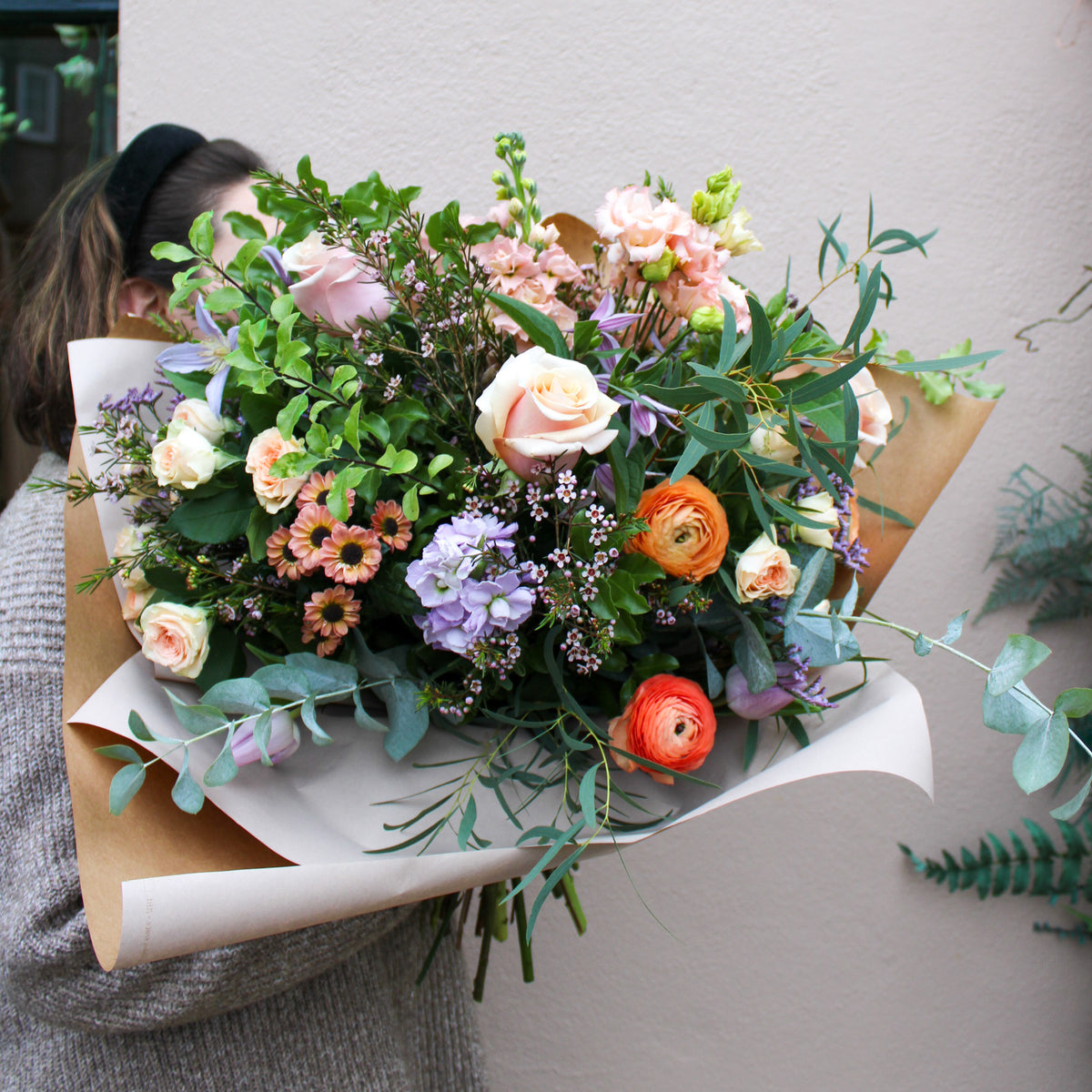 A person holds 'The Sunday' bouquet by Wild About Flowers, a large hand-tied arrangement with roses, ranunculus, daisies, lilacs, and assorted greenery wrapped in paper against a pale wall background.