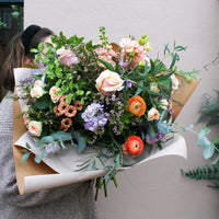 A person holds 'The Sunday' bouquet by Wild About Flowers, a large hand-tied arrangement with roses, ranunculus, daisies, lilacs, and assorted greenery wrapped in paper against a pale wall background.