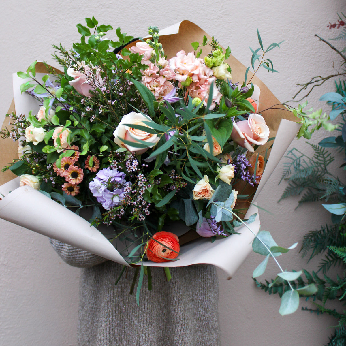 A person in a gray sweater holds 'The Sunday' hand-tied bouquet by Wild About Flowers, featuring assorted roses, greenery, and small blooms wrapped in white and brown paper against a light-colored wall.