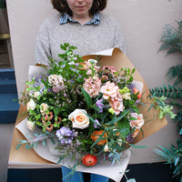 A person in a beige sweater holds 'The Sunday' hand-tied bouquet by Wild About Flowers, featuring roses, greenery, and pastel blooms wrapped in brown paper, standing before a light-colored wall.