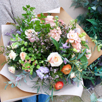 A person holds 'The Sunday' bouquet by Wild About Flowers, featuring pale pink roses, orange ranunculus, and vibrant greenery, all wrapped in brown and white paper—a fresh and stunning spring arrangement.