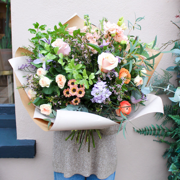 A person holds 'The Sunday' bouquet of Lilac and Peach flowers by Wild About Flowers, a hand-tied arrangement wrapped in brown paper with pink roses, purple and orange blooms, and greenery, set against a beige wall.