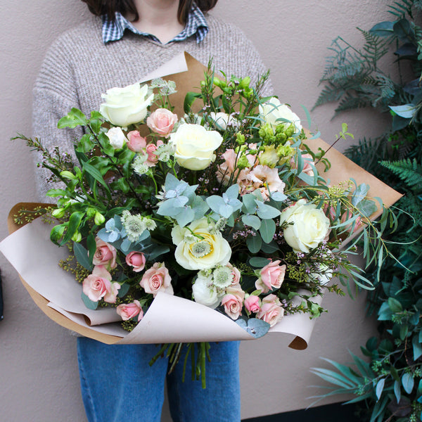 A person in a gray sweater and blue jeans holds the Wild About Flowers 'Sweet Blossom' Pink and White Fresh Flower Bouquet wrapped in brown paper. The background shows a beige wall and lush green plants.