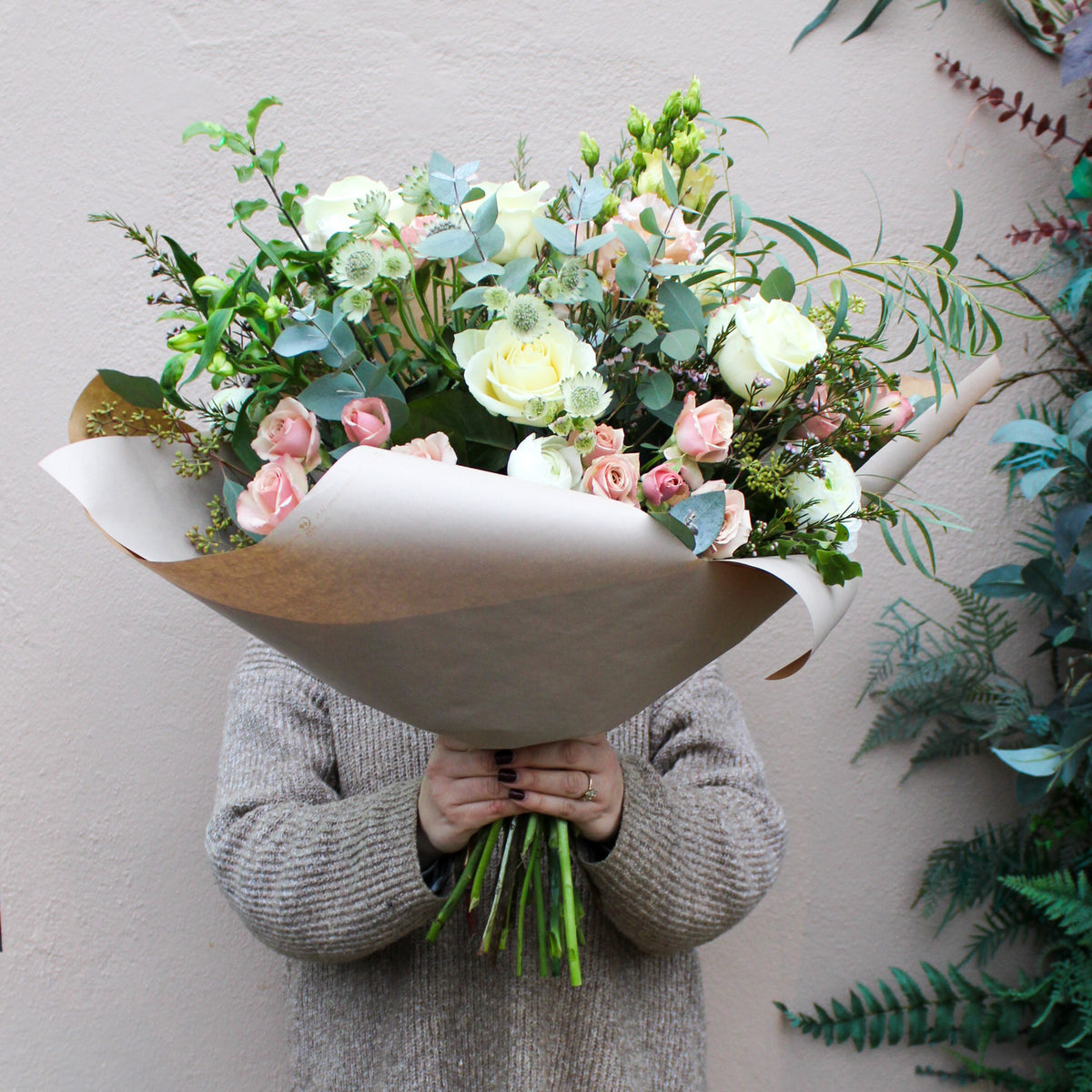 A person holds the 'Sweet Blossom' Pink and White Fresh Flower Bouquet by Wild About Flowers—premium white and pale pink roses with greenery, elegantly hand-tied and wrapped in brown paper—for Valentine's Day against a neutral wall.