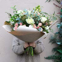 A person holds the 'Sweet Blossom' Pink and White Fresh Flower Bouquet by Wild About Flowers—premium white and pale pink roses with greenery, elegantly hand-tied and wrapped in brown paper—for Valentine's Day against a neutral wall.