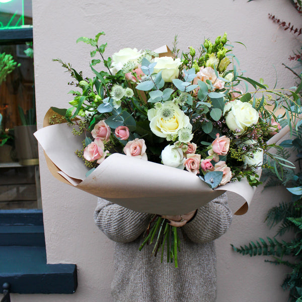 A person in a gray sweater holds the Wild About Flowers 'Sweet Blossom' Pink and White Fresh Flower Bouquet—a hand-tied arrangement—in front of their face, standing against a light-colored wall.