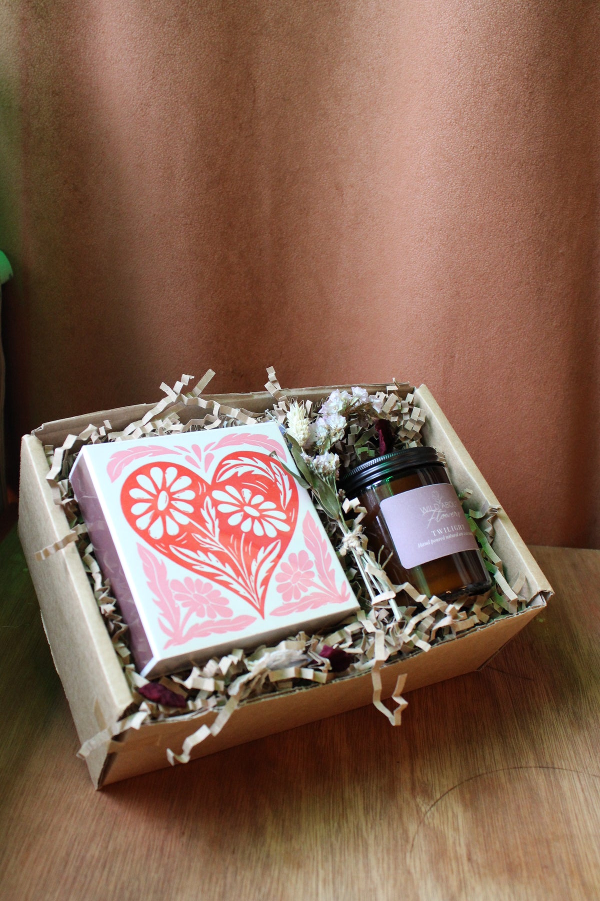 The 'The Perfect Match' Gift Box by Wild About Flowers features a card with a large red heart and floral designs, plus a natural soy wax candle in an amber jar, all presented with shredded paper filler in a stylish box.