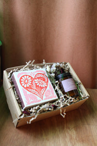 The 'The Perfect Match' Gift Box by Wild About Flowers features a card with a large red heart and floral designs, plus a natural soy wax candle in an amber jar, all presented with shredded paper filler in a stylish box.