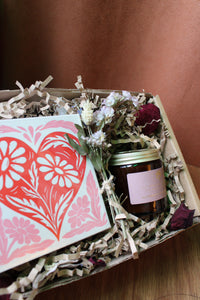 The Perfect Match Gift Box by Wild About Flowers features a natural soy wax candle, pink and orange heart card, dried flowers, and filler—displayed on wood with a brown fabric backdrop—perfect for Valentine’s Day.