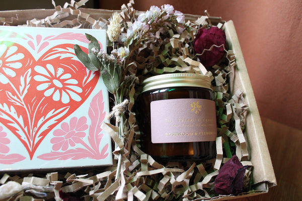 The Perfect Match Gift Box by Wild About Flowers includes a floral card with a red heart, Coconut & Geranium soy candle, dried flowers, and brown crinkle paper—an ideal and thoughtful Valentine's Day gift.