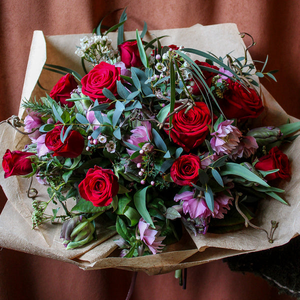 The 'True Romantic' Valentines Bouquet by Wild About Flowers features Red Naomi Roses, purple blooms, and greenery wrapped in brown paper—a perfect romantic floral arrangement.