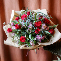 The 'True Romantic' Valentines Bouquet by Wild About Flowers features Red Naomi Roses and purple blooms with lush green foliage, wrapped in brown paper and shown against a brown fabric background.