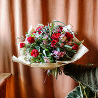 The 'True Romantic' Valentines Bouquet by Wild About Flowers features Red Naomi Roses and pink blooms, wrapped in brown paper—an ideal romantic arrangement, shown held by someone in a green sweater with a soft brown curtain and leafy plant behind.