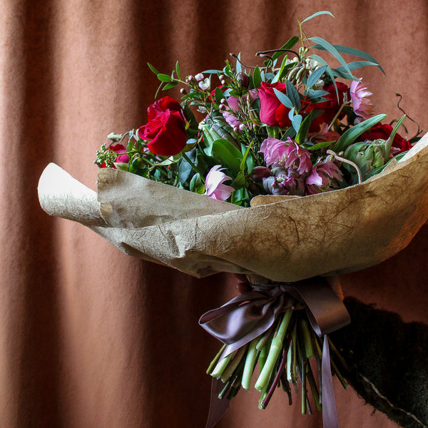 A hand holds the 'True Romantic' Valentines Bouquet by Wild About Flowers, with red and pink blooms and green leaves, wrapped in brown paper and tied with a purple ribbon, set before a brown curtain.