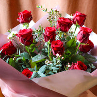 The Ultimate Valentine" Red Rose Bouquet by Wild About Flowers features luxurious red roses, green leaves, and white flowers in light pink wrap—an elegant Valentine’s Day gift.