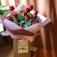 The Ultimate Valentine Red Rose Bouquet by Wild About Flowers features luxury red roses with greenery in pink wrap, displayed on a brown box before a curtain, and includes a card labeled "Wild About Flowers.