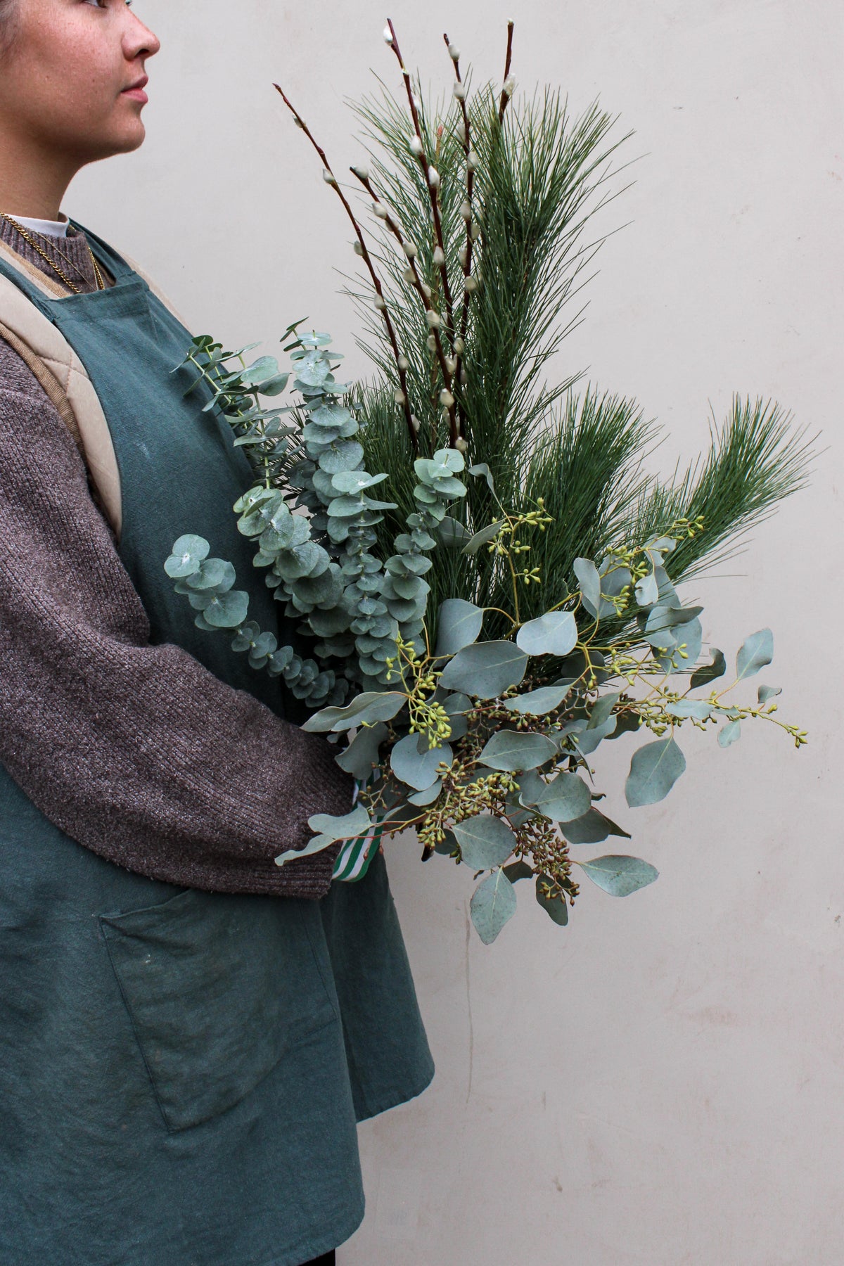 A person in a green apron holds Wild About Flowers' Wilder One Foliage Stems bouquet with eucalyptus, pine needles, and twigs against a light background.