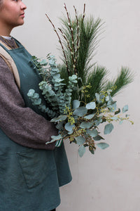 A person in a green apron holds Wild About Flowers' Wilder One Foliage Stems bouquet with eucalyptus, pine needles, and twigs against a light background.