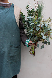 A person in a green apron and brown sweater holds the Wilder One Foliage Stems by Wild About Flowers, featuring festive eucalyptus and pine branches, against a plain light background.